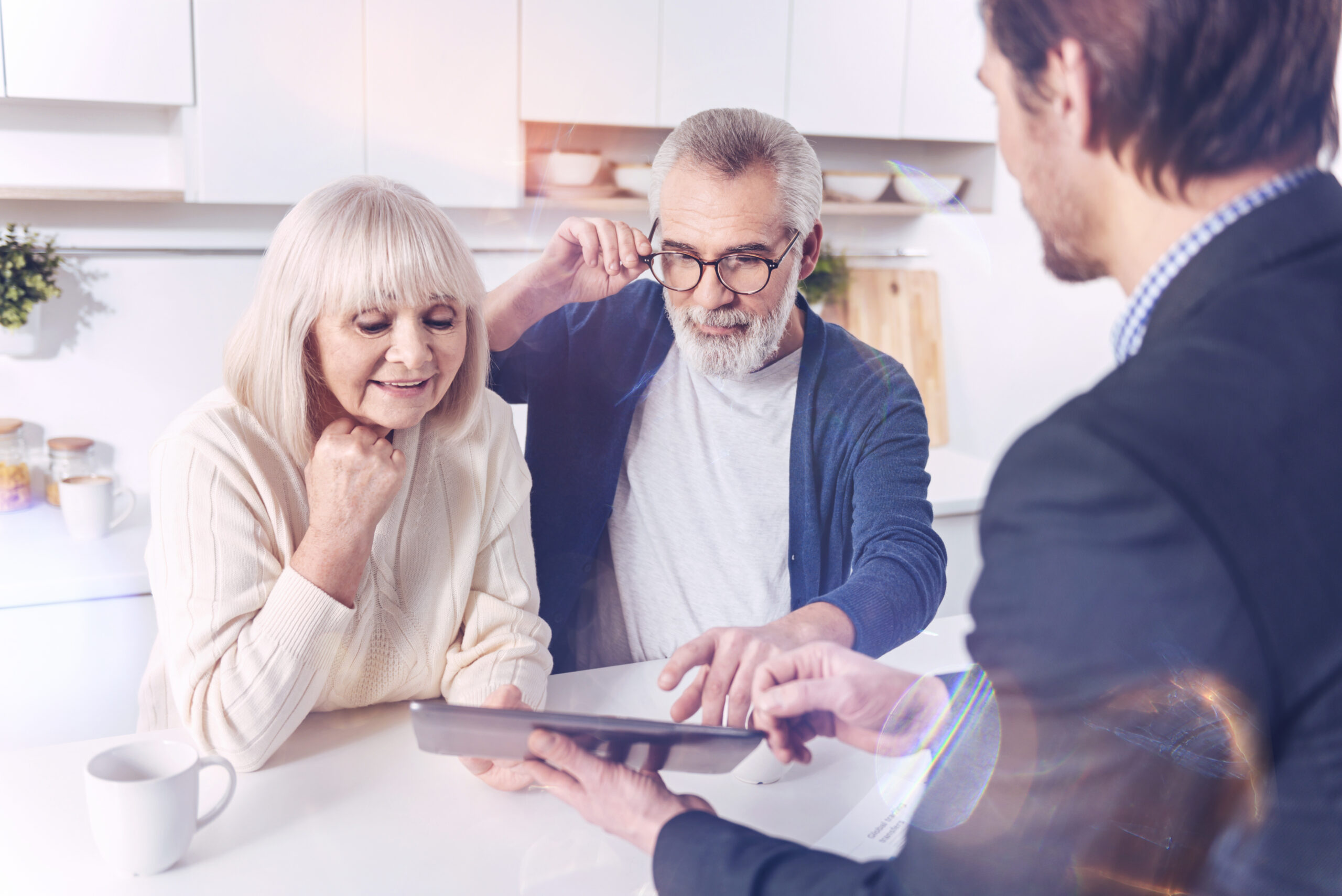 Just have a look. Positive elderly couple looking at the screen of tablet while talking with their real estate agent and meeting in the kitchen